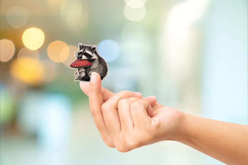 May include: A raccoon sticker with a red and white mushroom. The raccoon is black, gray, and white. The sticker is held between two fingers. The background is blurred with bokeh lights.
