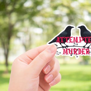 Puede incluir: Una pegatina con dos cuervos negros posados en una rama, con las palabras "Attempted Murder" en letras góticas rojas. La pegatina tiene un borde blanco y se sostiene sobre un fondo verde borroso.