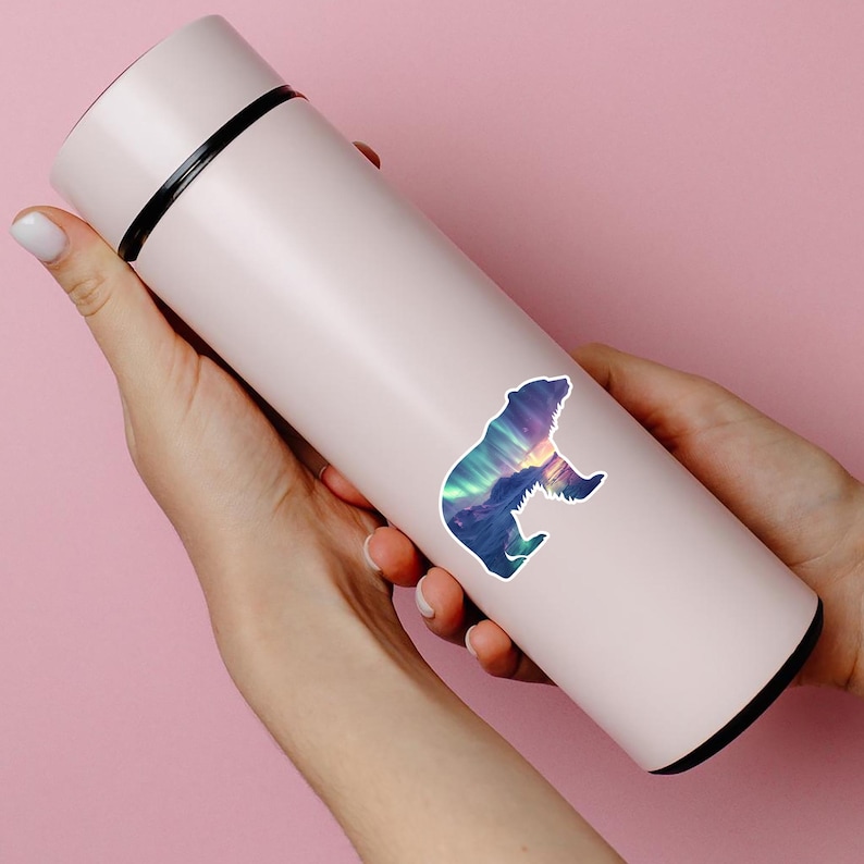 May include: A pale pink insulated water bottle with a black rim and a colourful bear sticker. The sticker shows a bear silhouette with a landscape of mountains and an aurora borealis design in blue, purple, and yellow.