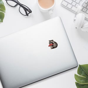 May include: A sticker featuring a raccoon wearing glasses and holding a red heart-shaped object. The sticker is on a silver laptop, surrounded by a white keyboard, headphones, and a cup of coffee.