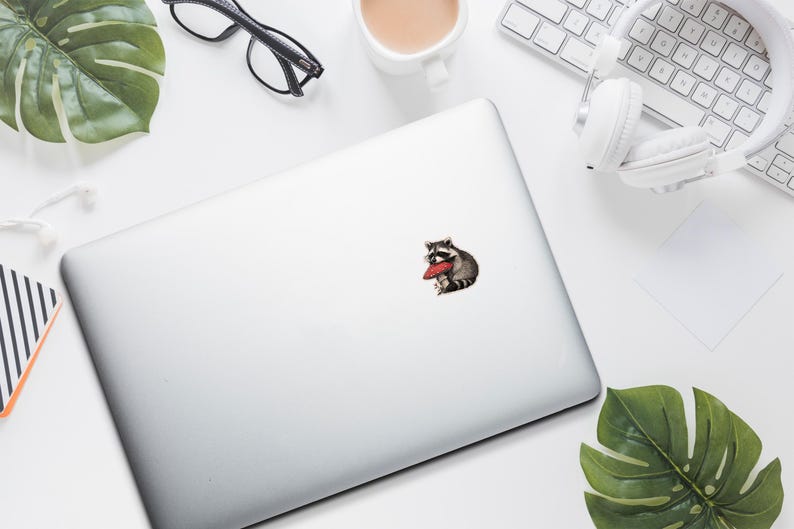 May include: A sticker featuring a raccoon wearing glasses and holding a red heart-shaped object. The sticker is on a silver laptop, surrounded by a white keyboard, headphones, and a cup of coffee.