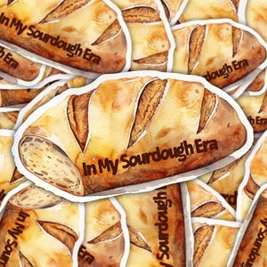 May include: A pile of stickers depicting a watercolour illustration of a sourdough loaf. The bread is golden brown with a textured crust and the text "In My Sourdough Era" in a dark brown font.