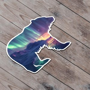 May include: A bear-shaped sticker with a white border, depicting a vibrant mountain landscape and aurora borealis. The sticker is set against a wooden background.