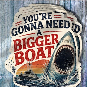 May include: A stack of die-cut stickers featuring a large shark emerging from the water next to a fishing boat. The text reads YOU'RE GONNA NEED A BIGGER BOAT in bold blue and red lettering over a sunset background with orange and yellow hues.