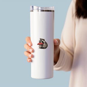 May include: White insulated tumbler featuring a whimsical illustration of a raccoon holding a red mushroom. The raccoon is depicted in shades of gray and black, with the mushroom in vibrant red and white. The tumbler has a silver lid.
