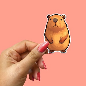 Pode incluir: Um adesivo de capivara de desenho animado marrom e branco segurado por uma m&atilde;o com esmalte de unha vermelho.