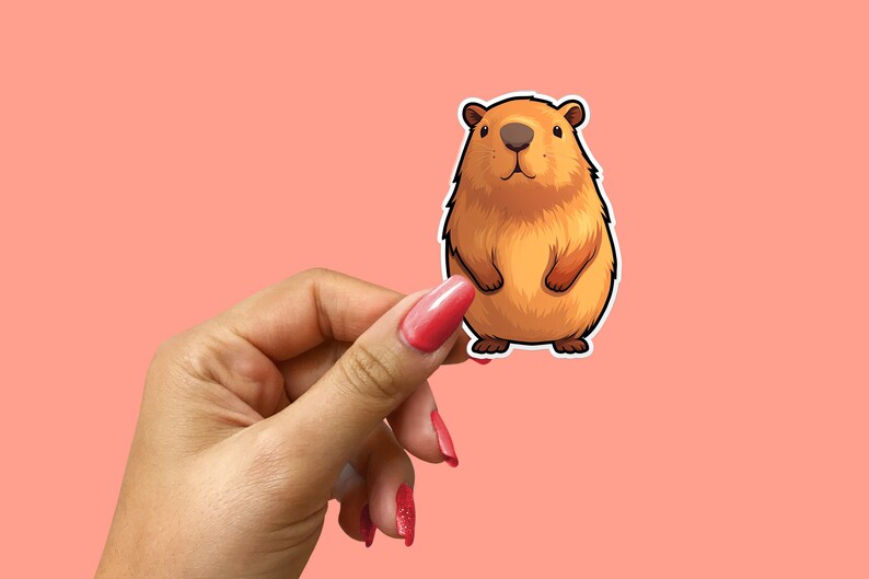 Pode incluir: Um adesivo de capivara de desenho animado marrom e branco segurado por uma m&atilde;o com esmalte de unha vermelho.