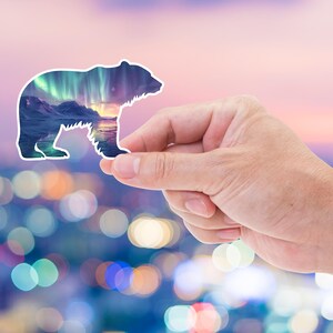 May include: A white-edged sticker shaped like a bear, featuring a landscape of mountains, water, and the aurora borealis. The sticker is held up against a blurred backdrop of city lights and a colourful sky.