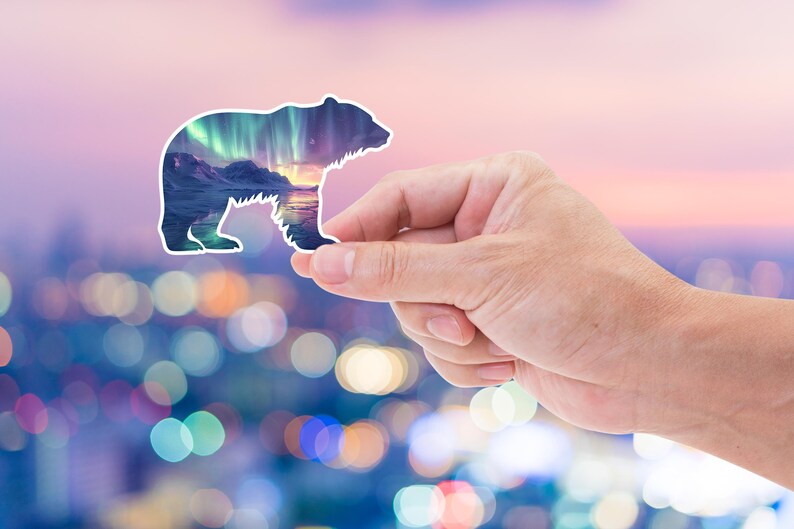 May include: A white-edged sticker shaped like a bear, featuring a landscape of mountains, water, and the aurora borealis. The sticker is held up against a blurred backdrop of city lights and a colourful sky.