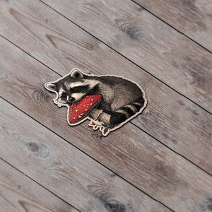 May include: A wooden sticker featuring a sleeping raccoon with a red mushroom. The raccoon is black, white, and gray, and the mushroom has white spots. The sticker is on a wooden surface.