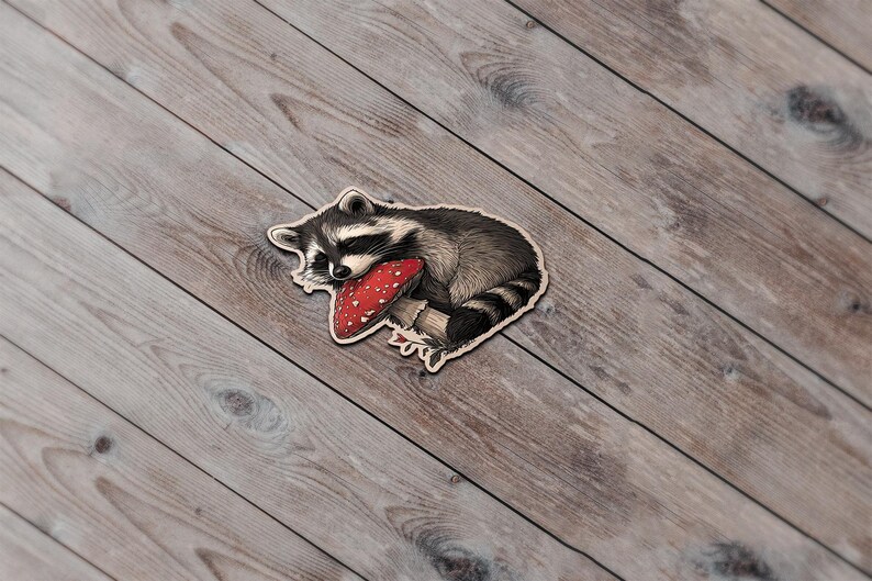 May include: A wooden sticker featuring a sleeping raccoon with a red mushroom. The raccoon is black, white, and gray, and the mushroom has white spots. The sticker is on a wooden surface.