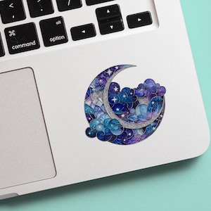 May include: A celestial-themed sticker featuring a crescent moon, clouds, and stars. The design incorporates shades of blue, purple, and silver, creating a dreamy, artistic aesthetic. The sticker is placed on a white laptop.