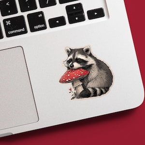 May include: A sticker featuring a raccoon holding a red mushroom with white spots. The raccoon is depicted in shades of gray and black, with a fluffy tail and a bandit mask. The mushroom has a white stem and is set against a white background.