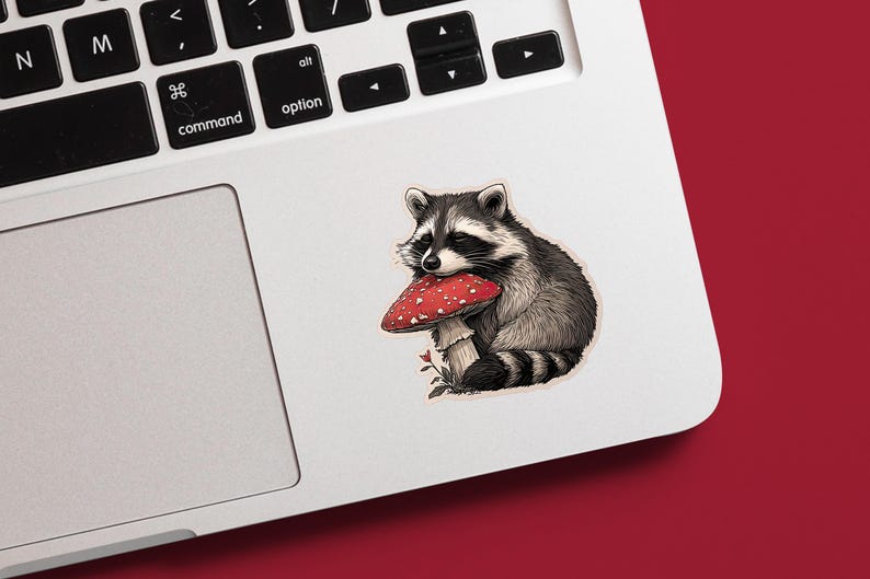 May include: A sticker featuring a raccoon holding a red mushroom with white spots. The raccoon is depicted in shades of gray and black, with a fluffy tail and a bandit mask. The mushroom has a white stem and is set against a white background.