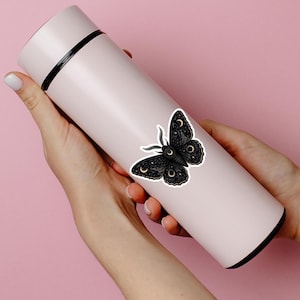 Puede incluir: Una botella de agua aislante cilíndrica de color rosa claro con una pegatina de polilla negra que presenta detalles de lunas crecientes y estrellas doradas. La botella tiene un borde negro en la parte superior e inferior, con un diseño sencillo.