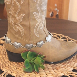 May include: A tan mid-calf boot featuring white decorative stitching on the shaft and a silver-toned chain accessory adorned with multiple heart-shaped charms wrapped around the ankle area of the footwear.