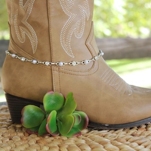 May include: A tan leather cowboy boot featuring white decorative stitching and a silver-toned chain anklet adorned with alternating small white pearls and clear round crystals wrapped around the lower ankle section of the boot.