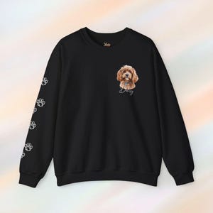 May include: Black sweatshirt featuring a watercolor portrait of a dog named Daisy. The dog is brown and white with a blue collar. White paw prints are printed down the left sleeve. The brand logo is in the upper right corner.