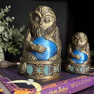 May include: Two bronze-colored figurines of a bearded creature with textured fur, each holding a smooth, bright blue sphere. The figures sit on circular bases decorated with teal and black circular patterns, resting on a Harry Potter book.