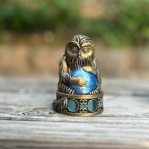 May include: A detailed, antique-style figurine of a monkey holding a blue orb. The monkey is bronze-colored with black accents, and the base features teal and black circular designs. The figurine is set against a blurred green background.