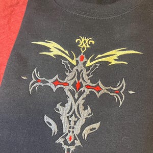 May include: Black sweatshirt with an embroidered cross design. The cross features red, gray, and yellow accents. The design is centered on the chest of the sweatshirt.