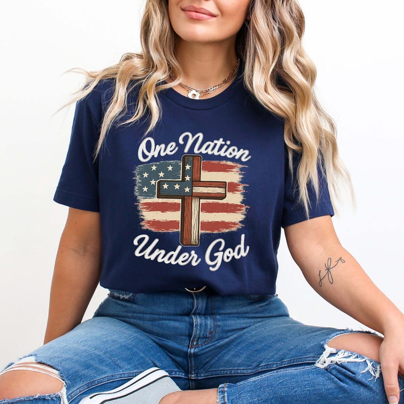 Pode incluir: T-shirt azul marinho com a frase "One Nation Under God" e um desenho com a bandeira americana e uma cruz. O design utiliza as cores vermelho, branco e azul, criando um tema patri&oacute;tico.