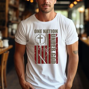 May include: White t-shirt with a distressed American flag design and the text "ONE NATION UNDER GOD." The flag includes a cross within a circle of stars. The shirt has a crew neck and short sleeves.