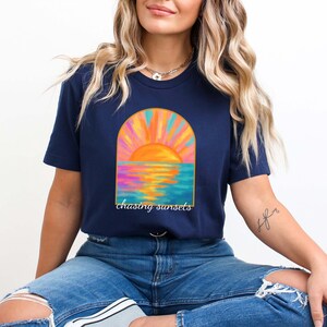 Puede incluir: Una camiseta azul marino con un gr&aacute;fico de una puesta de sol sobre el oc&eacute;ano en un marco arqueado. El texto "chasing sunsets" est&aacute; impreso debajo del gr&aacute;fico.
