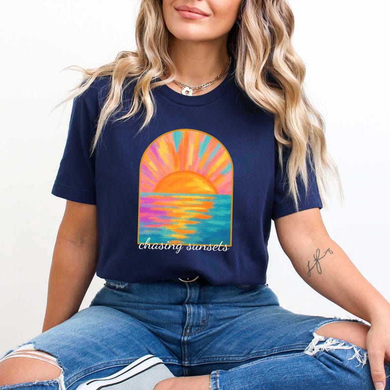 Puede incluir: Una camiseta azul marino con un gr&aacute;fico de una puesta de sol sobre el oc&eacute;ano en un marco arqueado. El texto "chasing sunsets" est&aacute; impreso debajo del gr&aacute;fico.