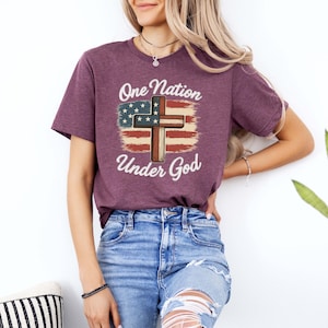 Pode incluir: Camiseta bord&ocirc; com a frase "One Nation Under God" acima de um desenho de cruz com um motivo de bandeira americana. O design incorpora as cores vermelho, branco e azul. A camisa &eacute; feita de um material macio.