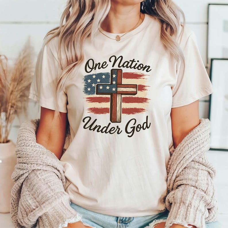 Pode incluir: Camiseta creme com um design patri&oacute;tico. O gr&aacute;fico inclui uma cruz sobreposta a uma bandeira americana estilizada com o texto "One Nation Under God" em uma fonte cl&aacute;ssica. A camiseta tem mangas curtas e gola redonda.