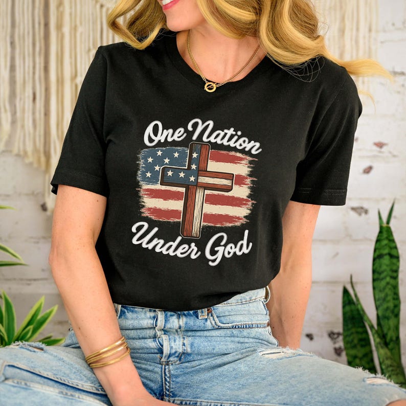 Pode incluir: T-shirt preto com a frase "One Nation Under God" acima de uma cruz e um desenho da bandeira americana. O design utiliza as cores vermelho, branco e azul. Uma pe&ccedil;a de roupa casual.