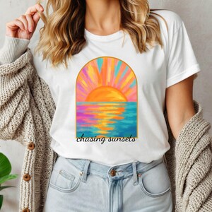 Puede incluir: Camiseta blanca con un gr&aacute;fico de puesta de sol colorido y el texto "chasing sunsets" impreso en la parte delantera.