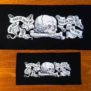 Könnte beinhalten: Zwei schwarze Stoffpatches mit weißem Text und Bildern. Der größere Patch zeigt einen Totenkopf mit Knochen und den Text "Das ist mein rot: Das end bedeckt. Wan alle ding beschlüßt der todt." Der kleinere Patch zeigt denselben Totenkopf mit Knochen und den Text "Das ist mein rot: Das end bedeckt. Wan alle ding beschlüßt der todt."