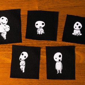 Puede incluir: Cinco parches de tela negra con ilustraciones blancas de espíritus Kodama. Cada parche presenta una pose diferente de los espíritus del bosque, con variaciones en su postura e interacción. Los parches están dispuestos sobre una superficie de madera.