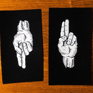 Könnte beinhalten: Zwei schwarze Stoffpatches mit weißen gestickten Handgesten. Der linke Patch zeigt eine Hand mit zwei ausgestreckten Fingern, der rechte Patch zeigt eine Hand mit einem ausgestreckten Finger.