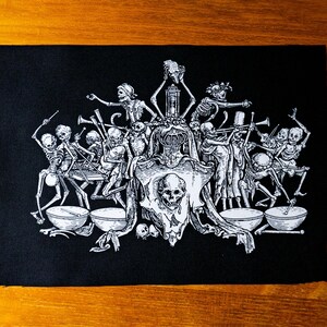 May include: Black and white embroidered patch depicting a group of skeletons playing musical instruments. The skeletons are arranged in a circle around a skull wearing a crown. The patch is on a black background.