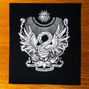 Op de afbeelding: Zwart-wit geborduurde patch met een gestileerde feniks met uitgespreide vleugels, een zon erboven en de tekst "utam aeternam uiuo ut".