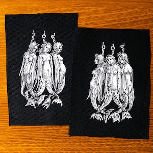 May include: Three black and white mermaids are hanging from chains on a black fabric patch. The mermaids are depicted in a vintage style with detailed features and flowing tails.