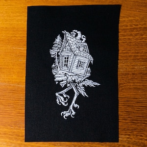 Puede incluir: Parche negro con una ilustración blanca de una casa con chimenea, posada sobre patas de pájaro. La casa tiene ventanas, una puerta y un árbol. El parche es rectangular.