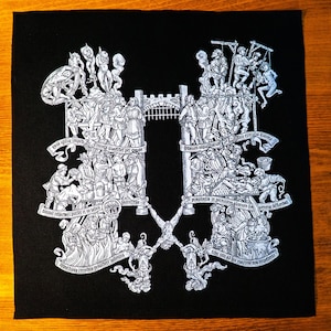 May include: Black square fabric with a detailed, white ink illustration. The artwork depicts a medieval scene with figures, structures, and banners. The design is symmetrical, with a central gate and text.