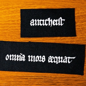 Könnte beinhalten: Zwei schwarze Stoffpatches mit weißem Text. Der obere Patch sagt "Anarchist" und der untere Patch sagt "omnia mors aequat".