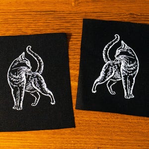 Op de afbeelding: Twee zwarte stoffen patches met een witte illustratie van een kat. De kat is in profiel afgebeeld, achterom kijkend met zijn staart omhoog. De patches zijn rechthoekig en liggen op een houten oppervlak.