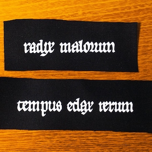 May include: Two black fabric labels with white text. The top label reads "radyr malovum" and the bottom label reads "tempus edax rerum".