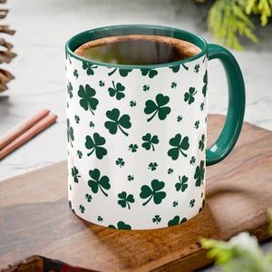 May include: A white ceramic mug with a green interior and handle, decorated with a pattern of green shamrocks. The mug is filled with a dark liquid, possibly coffee or tea, and sits on a wooden cutting board.