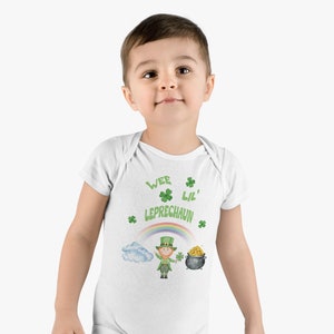 Puede incluir: Body de bebé blanco con el texto "Wee Lil' Leprechaun" en verde, junto con tréboles, un arcoíris, una nube, un duende y una olla de oro. El body es de manga corta.