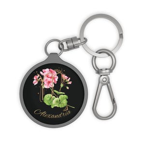 May include: A round keychain with a black background featuring a watercolor illustration of pink geranium flowers and green leaves. The word "Alexandria" is written in gold script. The keychain has a silver-colored metal ring and clasp.