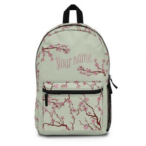 Puede incluir: Una mochila de color verde salvia claro con un patrón de ramas de cerezo de color marrón oscuro y flores rosas. El frente incluye un bolsillo con cremallera y el texto "Your name" en cursiva. Tiene un asa superior negra y un bolsillo lateral de malla.