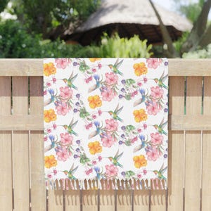 May include: A white rectangular towel featuring a repeating watercolor print of green and orange hummingbirds, pink hibiscus flowers, yellow blossoms, and small purple floral clusters. The bottom edge is finished with a row of white fringe.