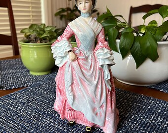 以下が含まれることがあります： ピンクと白のドレスを着た女性の人形。大きな白い帽子をかぶっています。青と茶色の模様の表面に立っています。